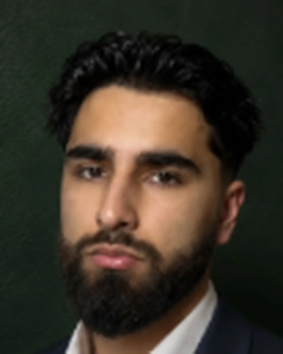 Anosh Mian Khel, founder of Sharp Studio, in a navy suit and white shirt against a dark green wall.